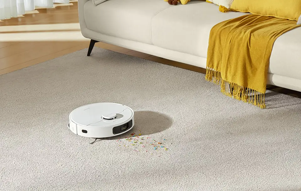 biały robot sprzątający odkurza dywan, w tle jasna kanapa i żółty koc