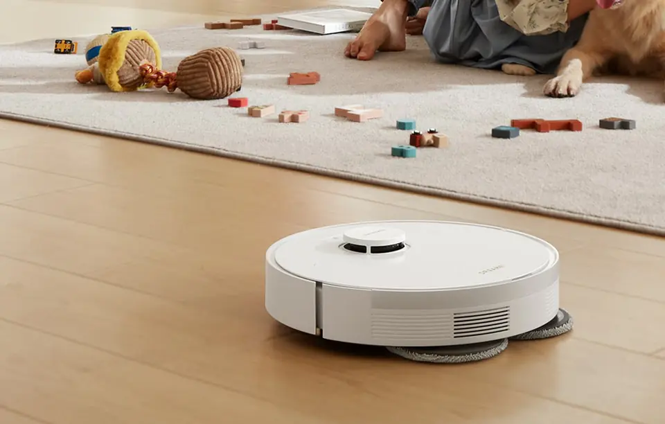 biały robot sprzątający mopuje drewnianą podłogę, w tle dywan z porozrzucanymi zabawkami