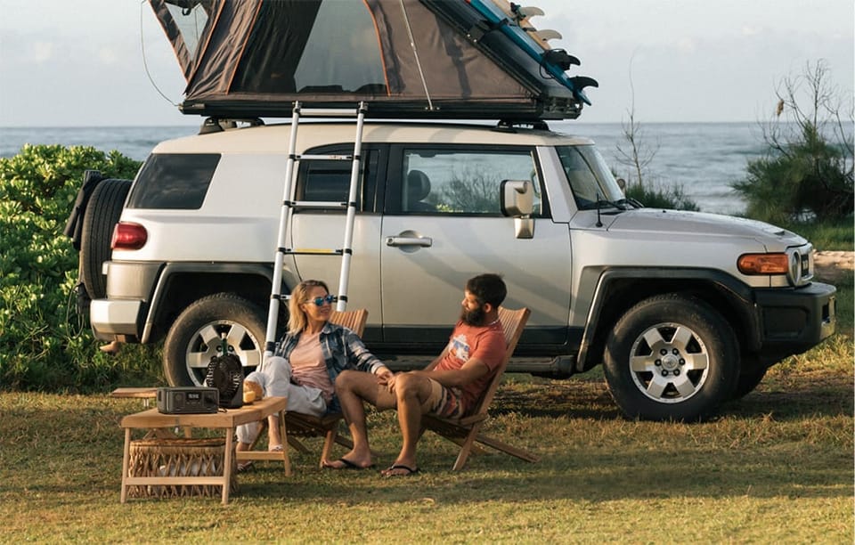 Para siedzi na krzesłach kempingowych obok samochodu terenowego z namiotem dachowym, na stole stoi stacja EcoFlow RIVER 3