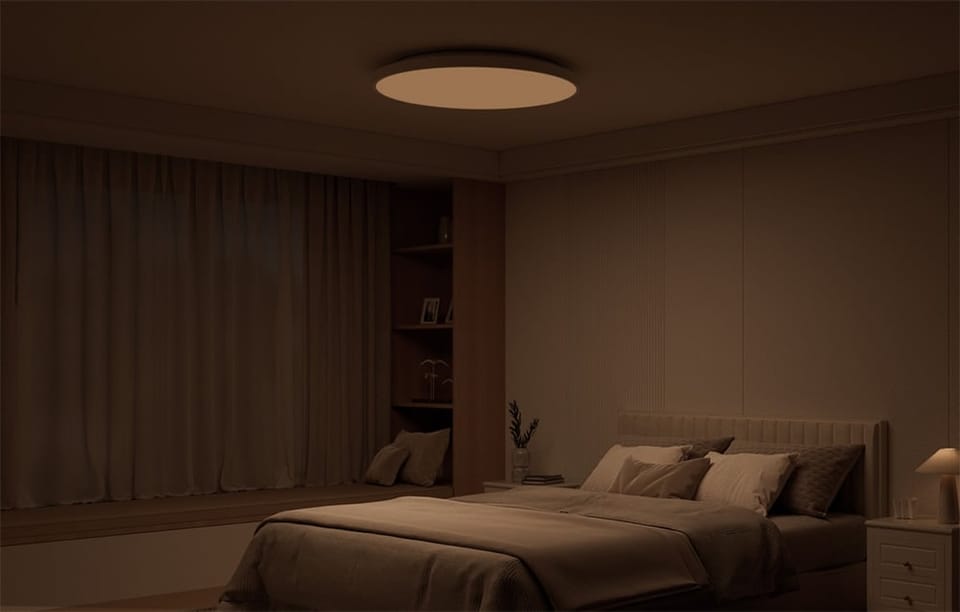 Przyciemnione wnętrze sypialni z łóżkiem i świecącą w ciepłym świetle inteligentną lampą sufitową Yeelight Mercury C300.