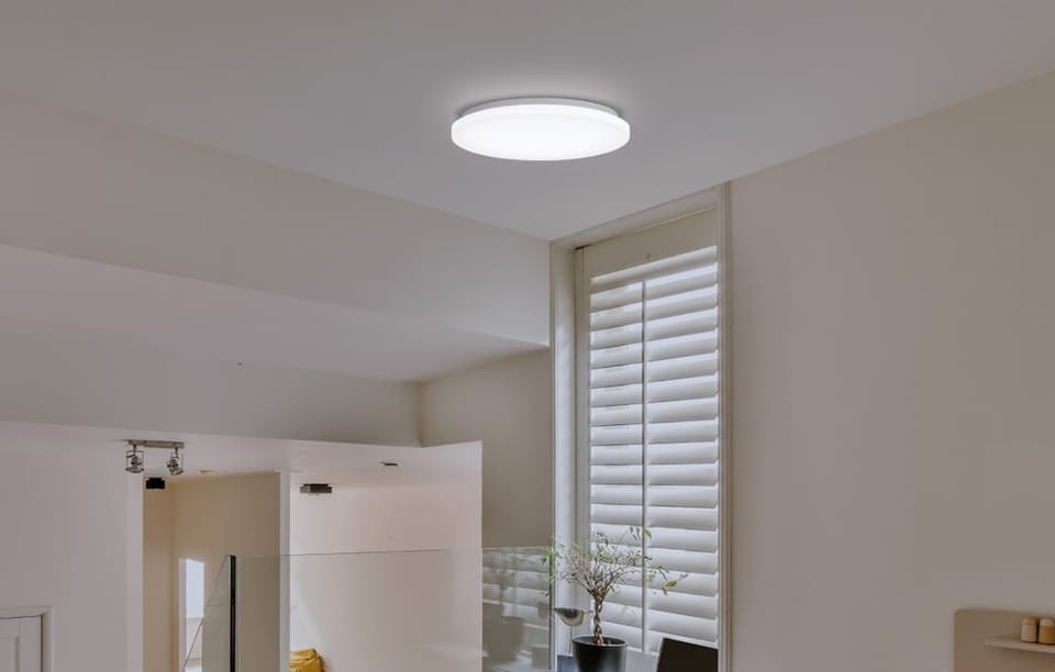 Lampa Yeelight MercuryE C300 zamontowana we wnętrzu z żaluzjami pionowymi i doniczką na parapecie.