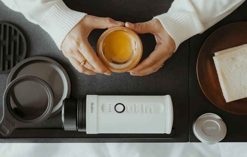 Kobieta trzyma w dłoni kubek z kawą. Obok na stoliku leży ekspres do kawy Outin.