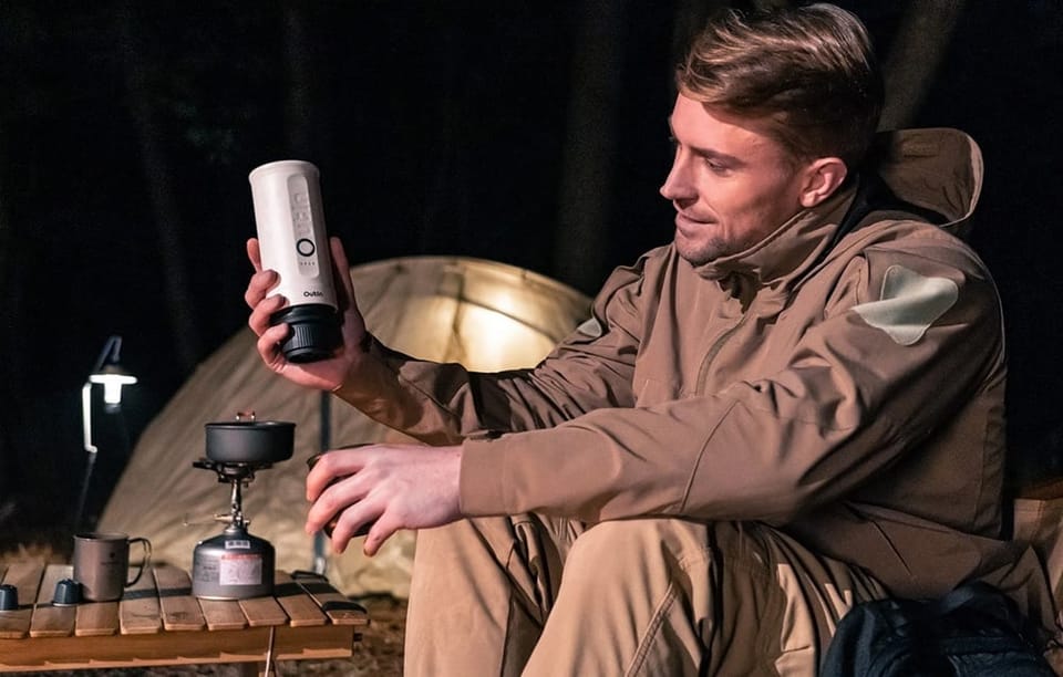 Mężczyzna w brązowej kurtce parzy kawę za pomocą bezprzewodowego ekspresu do kawy Outin. W tle rozbity namiot.