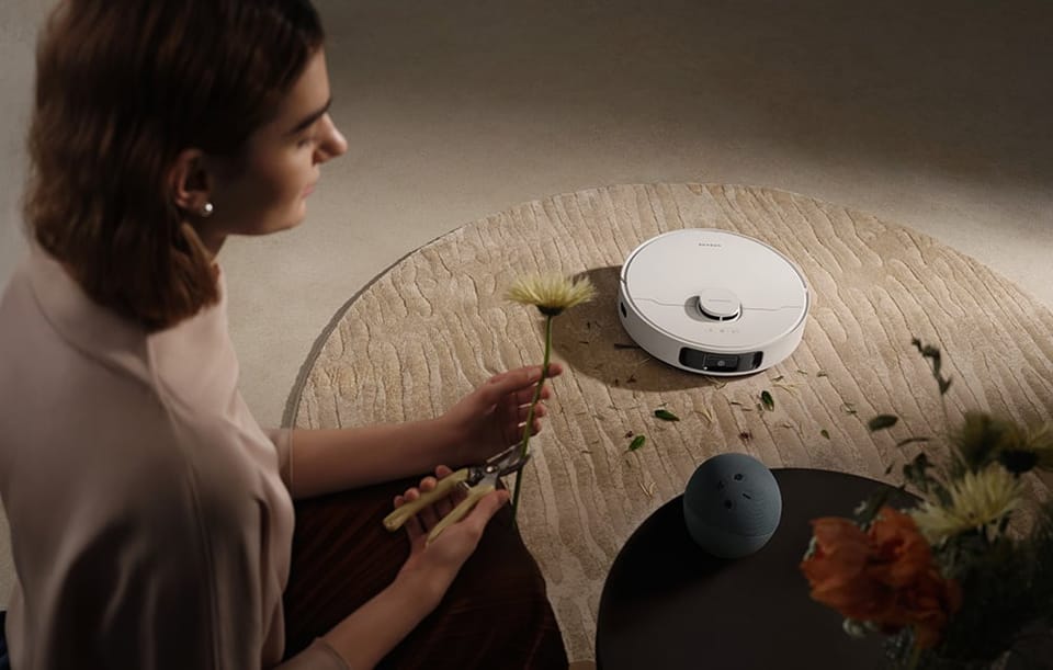 Kobieta przy okrągłym stole przycina kwiaty, a obok na dywanie jeździ robot sprzątający.
