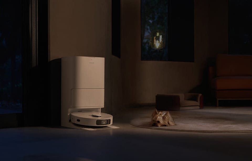 Robot sprzątający stoi w stacji dokującej wieczorem, a obok na dywanie leży mały pies.