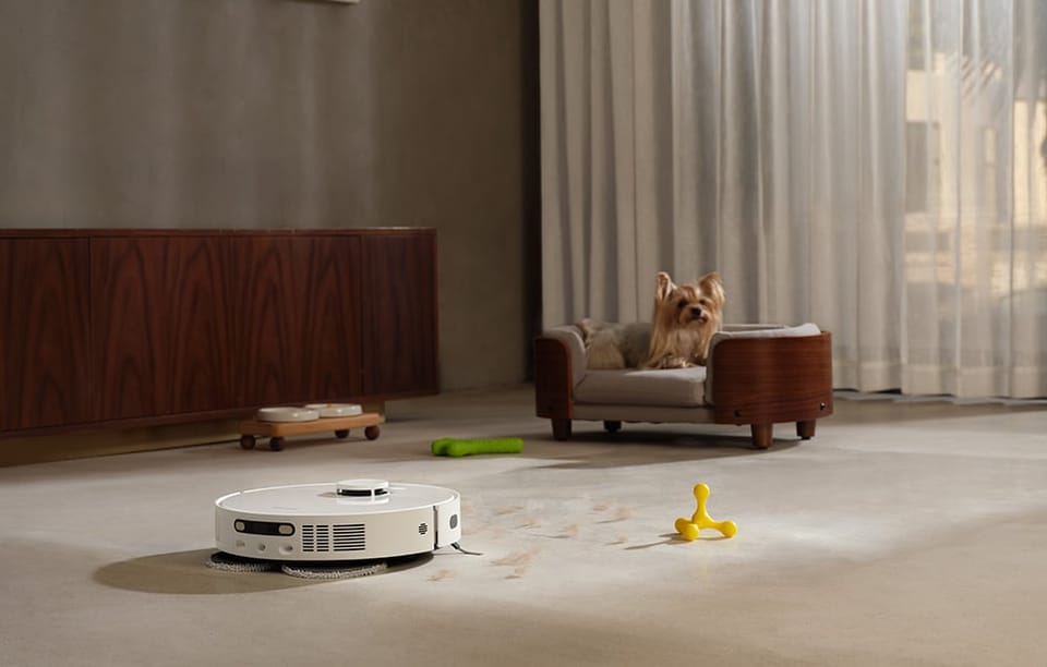 Robot sprzątający odkurza podłogę w pokoju, gdzie na kanapie leży mały pies; wokół zabawki.