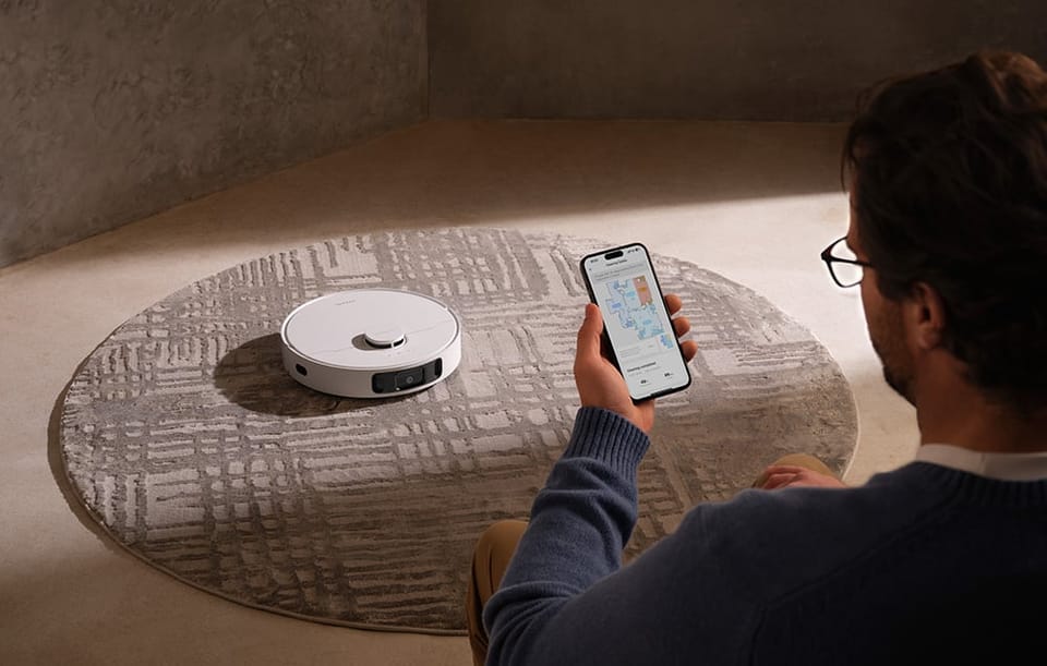 Mężczyzna siedzi na podłodze i patrzy na mapę sprzątania w aplikacji na smartfonie obok robota sprzątającego na okrągłym dywanie.