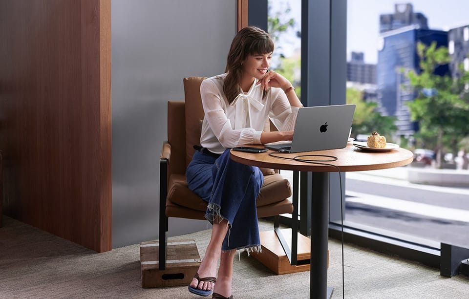 Kobieta w białej koszuli pracuje na MacBooku przy stoliku w kawiarni z widokiem na miasto