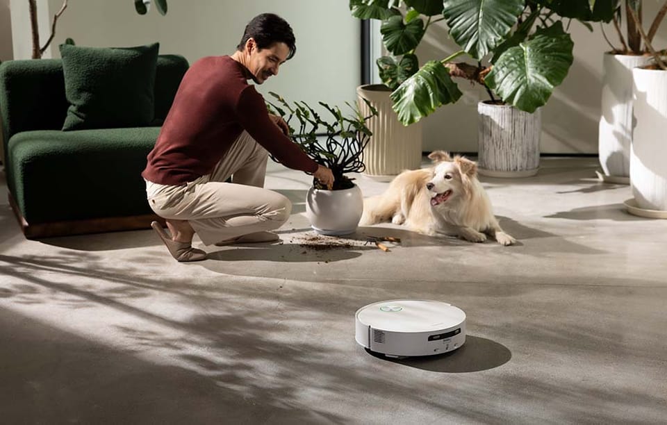 Mężczyzna przesadza roślinę w donicy, a obok na podłodze leży pies i robot sprzątający