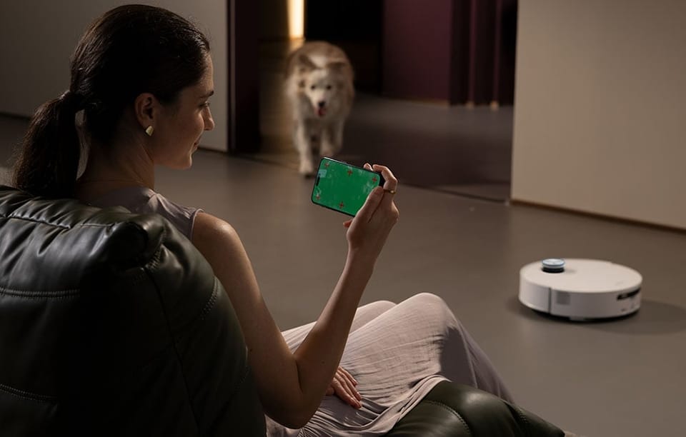 Kobieta siedzi na fotelu i steruje robotem sprzątającym za pomocą aplikacji na smartfonie, w tle pies