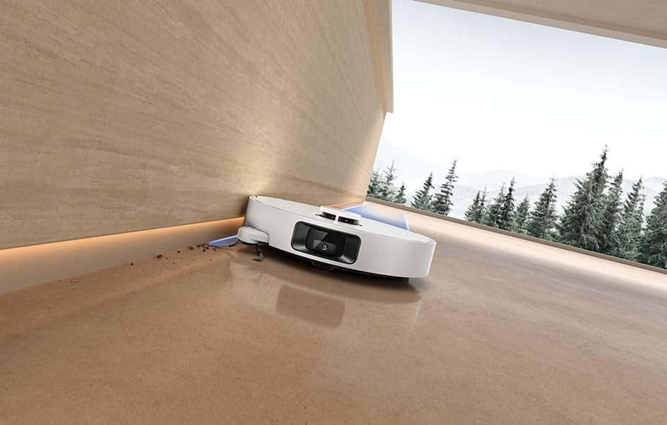 Robot Dreame D20 Pro jedzie wzdłuż ściany, zbierając zabrudzenia z podłogi przy krawędzi; za oknem widać drzewa.