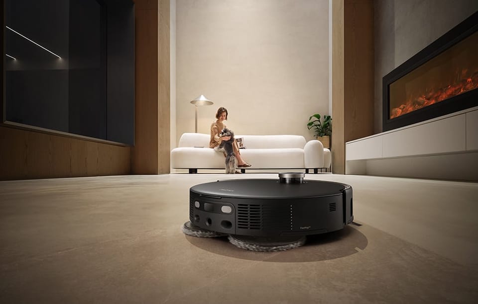 Kobieta siedzi na kanapie z psem. Z przodu robot sprzątający mopuje podłogę.