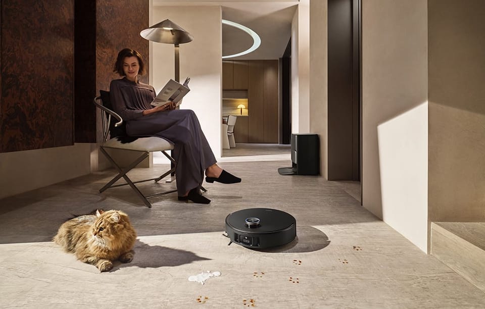 Kobieta siedzi na krześle i trzyma w dłoniach gazetę. Na podłodze znajdują się robot sprzątający i kot.