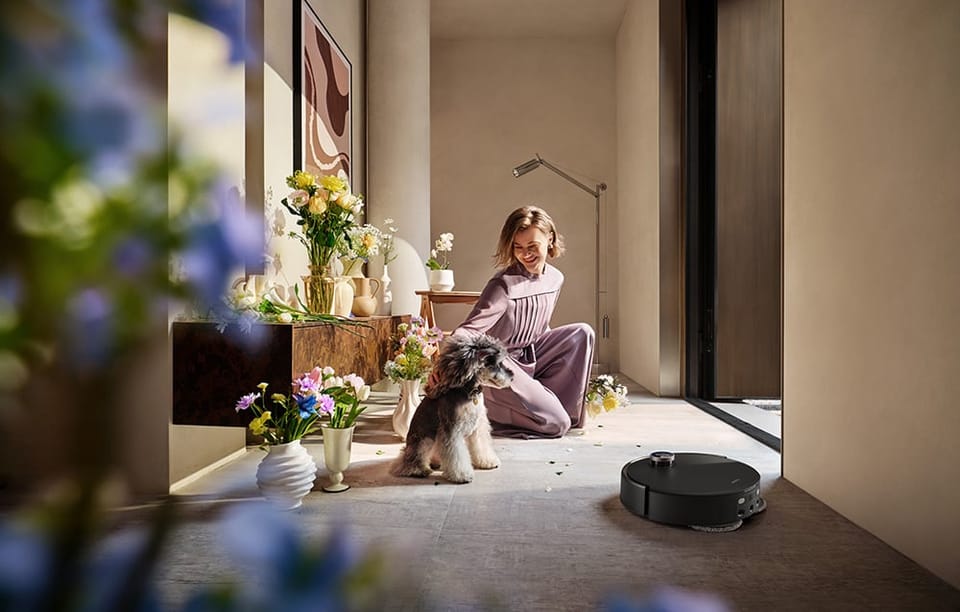 Kobieta uśmiecha się i głaszcze psa. Obok robot sprzątajay odkurza kafelki.