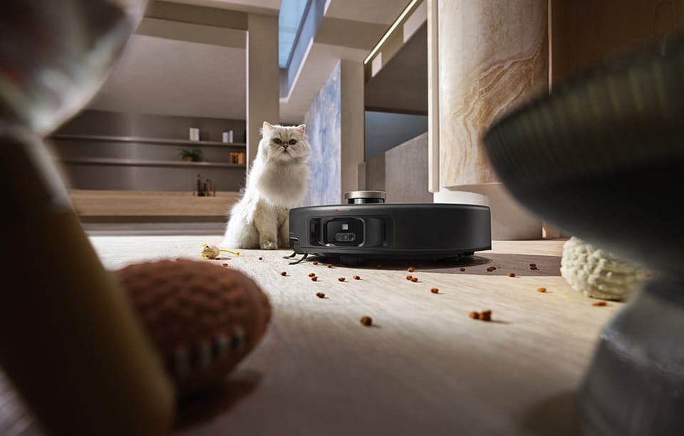Robot sprzątający zbiera rozsypaną na podłodze karmę. Za nim siedzi kot.