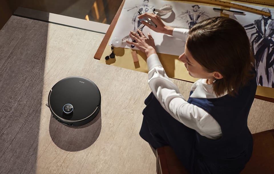 Kobieta siedzi przy stole i rysuje. Robot sprzątający odkurza podłogę.