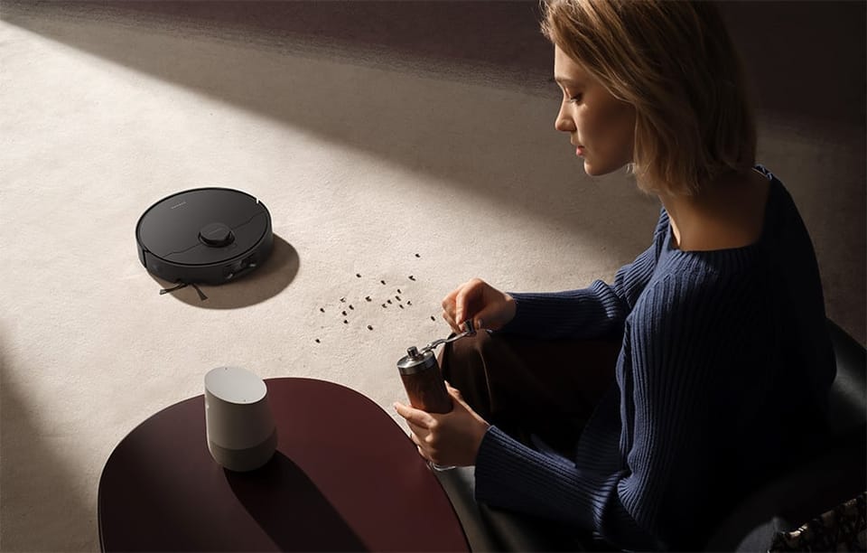 Kobieta siedzi na krześle przy stoliku i mieli kawę ręcznym młynkiem; na jasnym dywanie widać rozsypane drobiny, a w tle robot sprzątający Dreame D20 Pro Plus zbliża się, aby je posprzątać. 