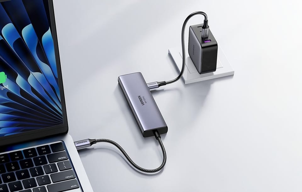 Hub UGREEN podłączony do laptopa i zasilacza USB-C, leżący na biurku obok pracującego MacBooka.