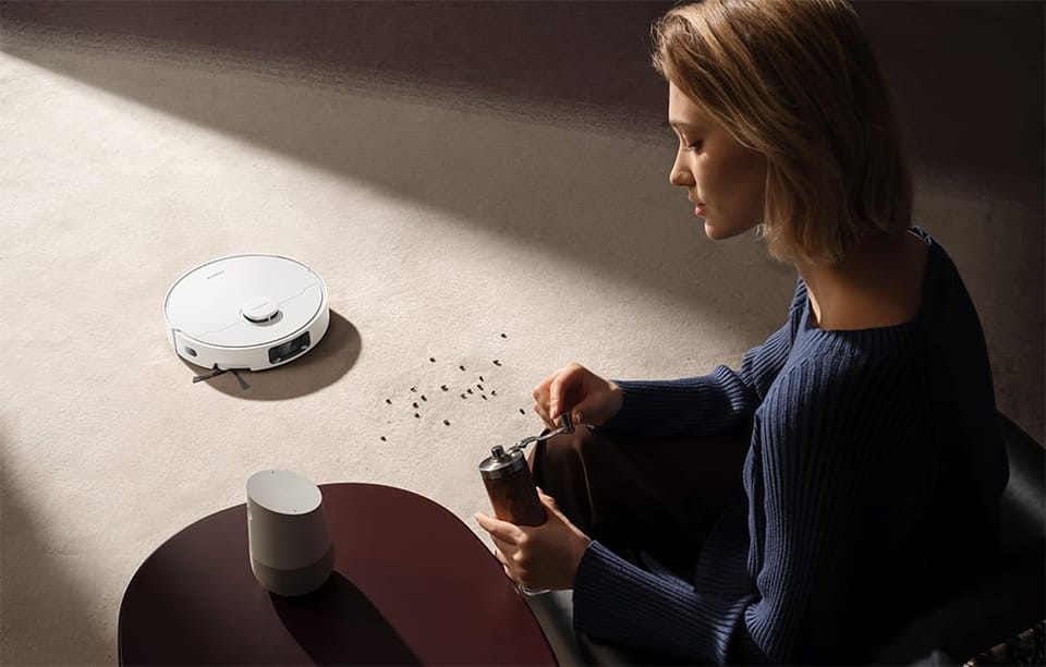 Kobieta siedzi na podłodze i mieli kawę, a obok na dywanie leżą rozsypane drobinki kawy; w jej kierunku jedzie biały robot sprzątający Dreame D20 Pro.