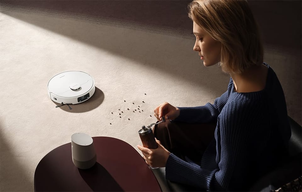 Kobieta z blond włosami siedzi na podłodze przy stoliku; obok niej robot sprzątający Dreame D20 Pro Plus odkurza rozsypane drobinki.