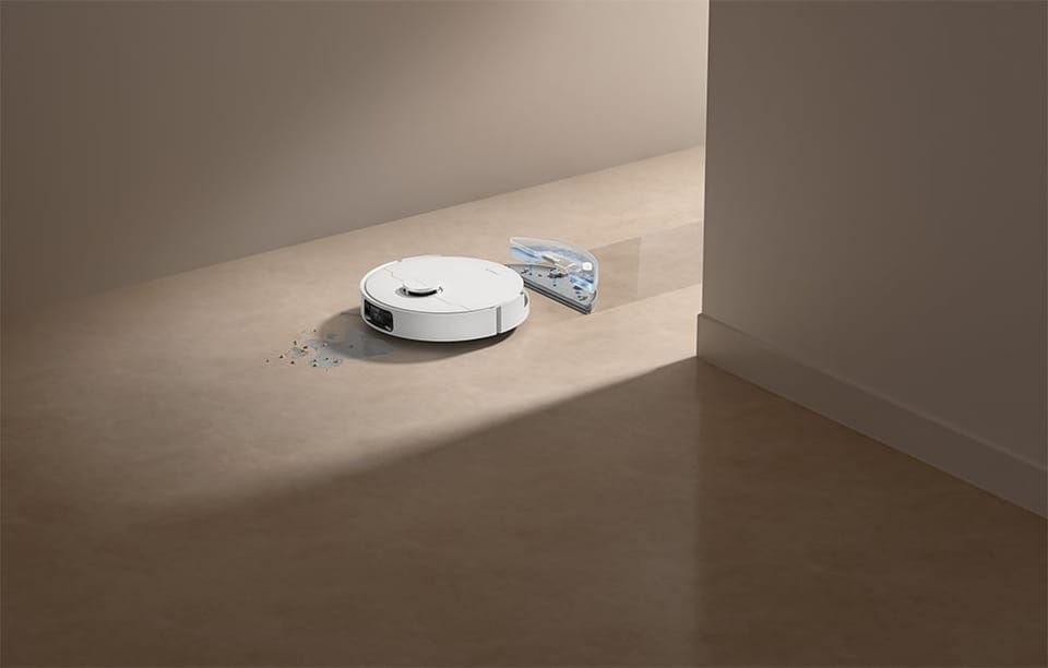 Przed robotem leżą rozsypane drobinki i okruchy, a obok widać przezroczystą, niebieską grafikę w kształcie wycinka koła z drobinkami i kroplami, unoszącą się nad podłogą.