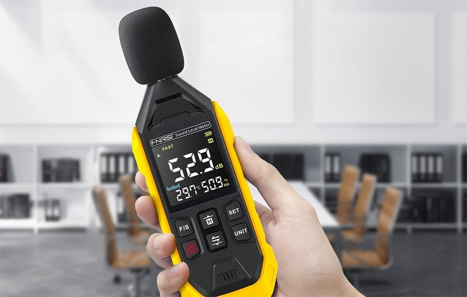 Ręka trzyma miernik poziomu dźwięku z osłoną mikrofonu i wyświetlaczem pokazującym 52,9 dB w biurze.