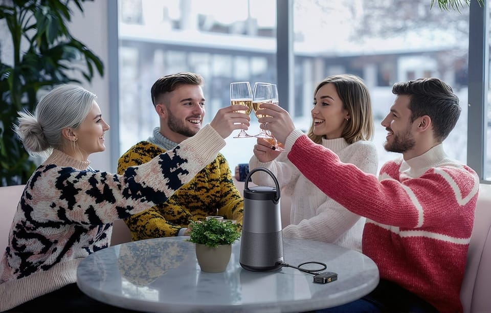 Czworo przyjaciół wznosi toast kieliszkami przy stole z głośnikiem i adapterem Bluetooth w jasnym pokoju