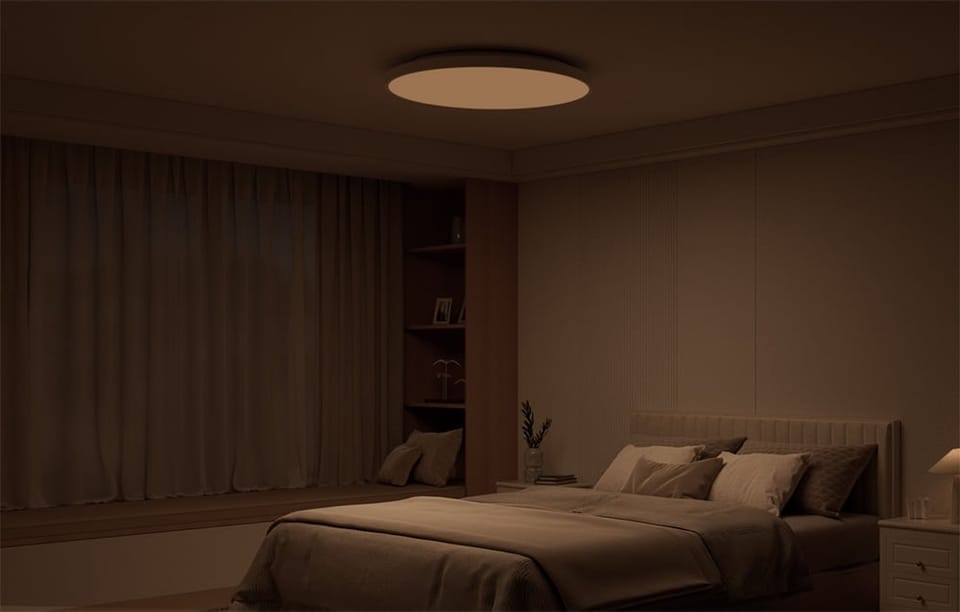 Przyciemnione wnętrze sypialni z łóżkiem i świecącą w ciepłym świetle lampą sufitową Yeelight Mercury C300.