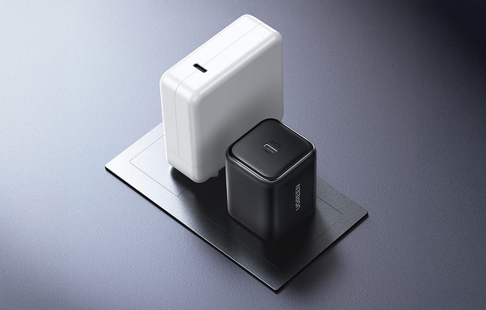 Czarna ładowarka UGREEN ustawiona obok większej białej ładowarki dla porównania rozmiaru na szarej powierzchni.