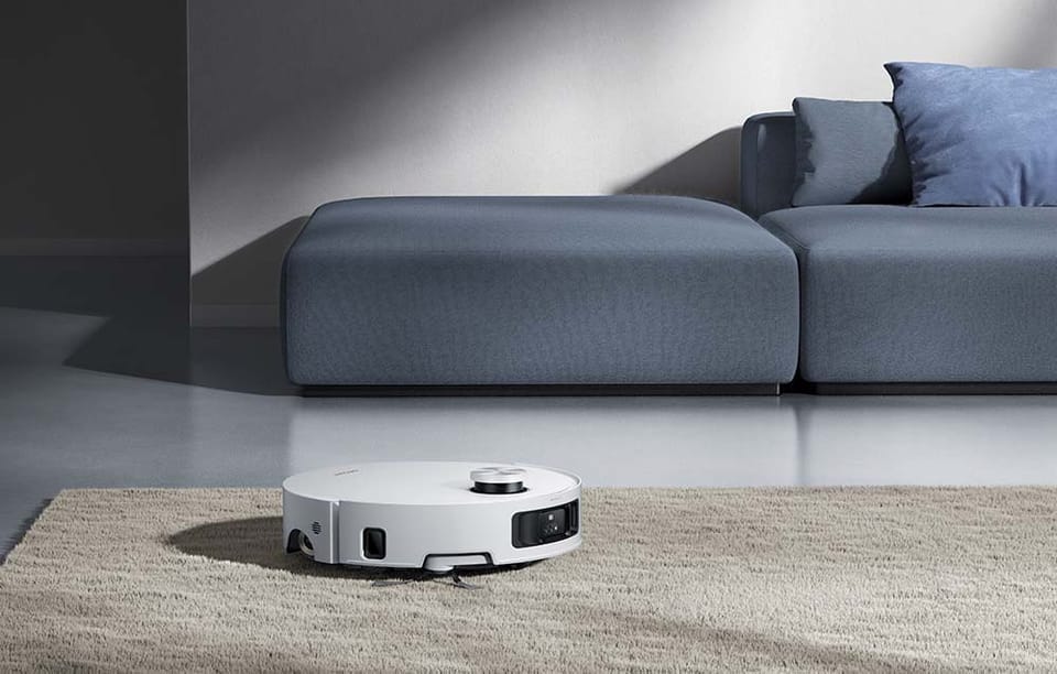 Biały robot sprzątający odkurza dywan w salonie. Za nim szara kanapa z poduszkami.