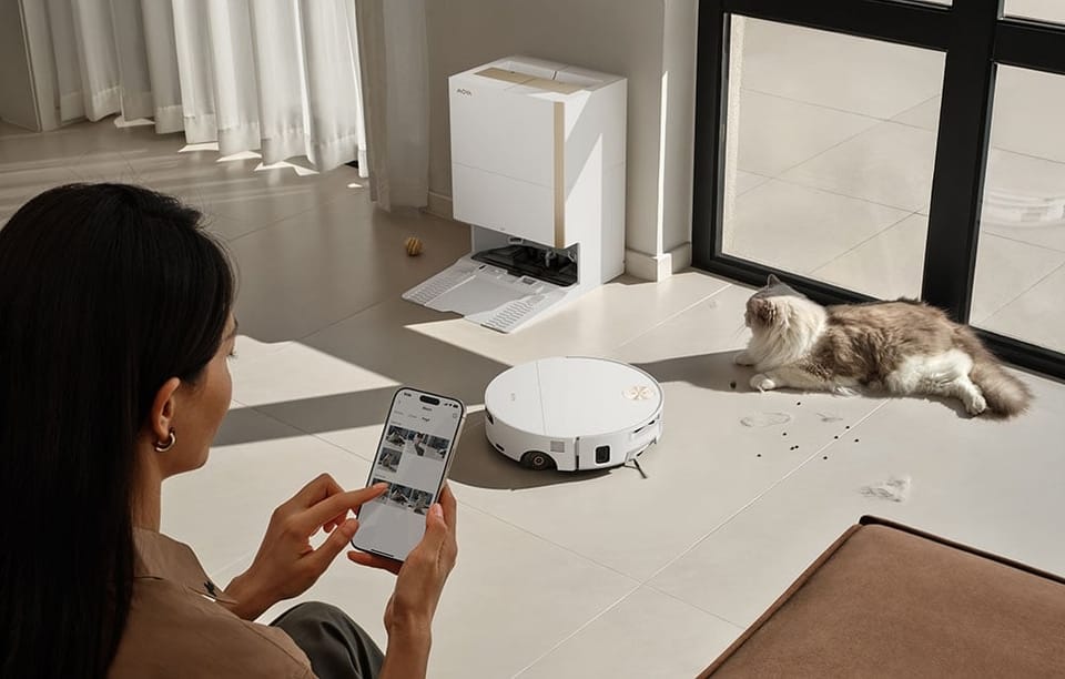 Kobieta trzyma telefon z aplikacją, obok biały robot sprzątający, stacja dokująca MOVA i leżący kot