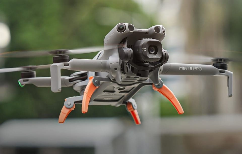Latający dron DJI Mini 5 Pro z rozłożonymi pomarańczowymi nogami, unoszący się w powietrzu