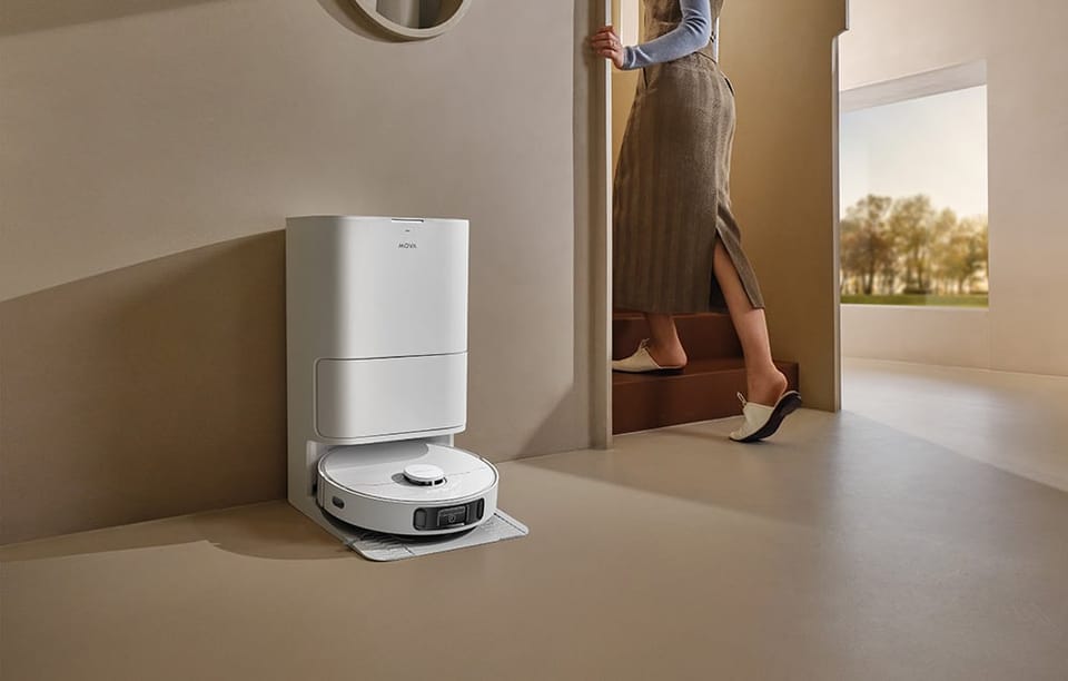 Kobieta wychodzi z pomieszczenia. Pod ścianą znajduje się robot sprzątający umieszczony w stacji dokującej,