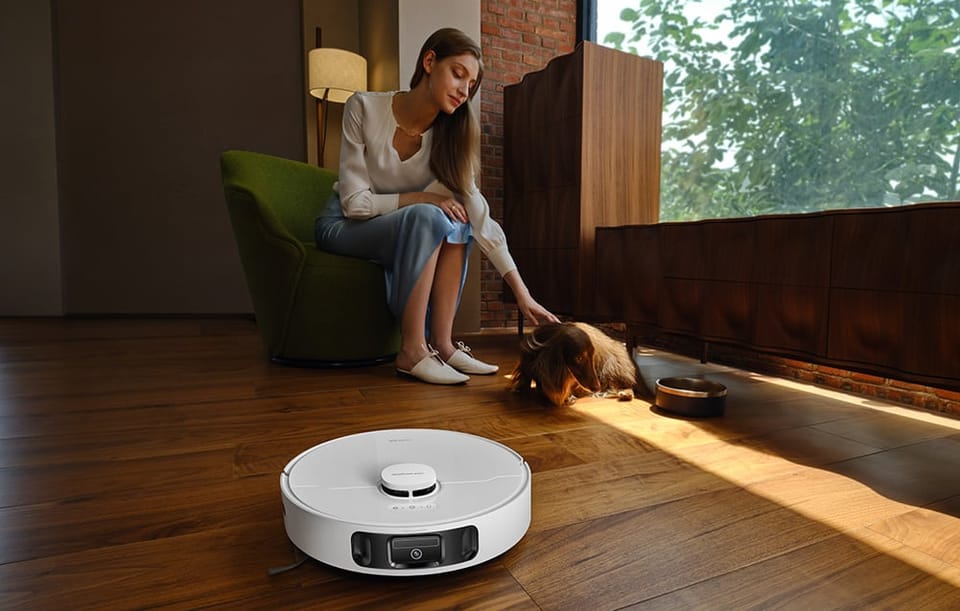 Kobieta siedzi na fotelu i głaszcze psa. Z przodu robot sprzątający odkurza podłogę. 