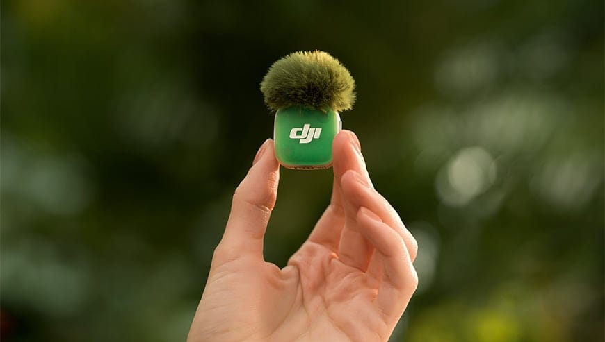 Dłoń unosi zielony mikrofon DJI Mic Mini 2 z nałożoną na niego futrzastą osłoną przeciwwietrzną na tle roślinności
