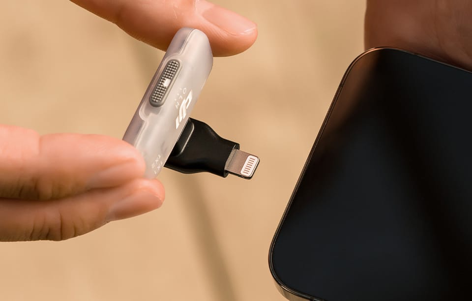 Dłonie trzymające biały odbiornik DJI z wpiętym czarnym adapterem Lightning przy krawędzi smartfona