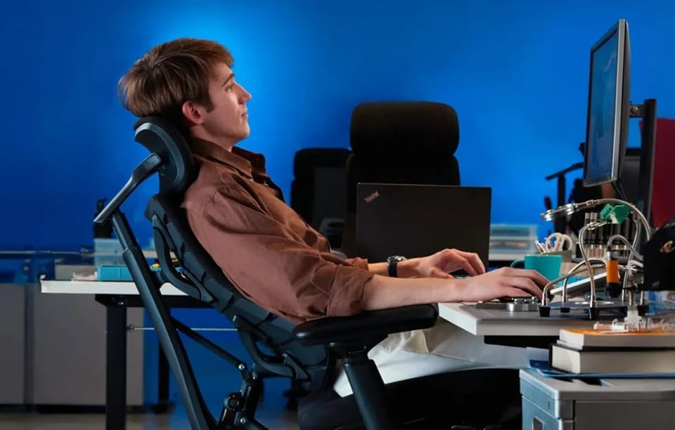 Mężczyzna siedzący na ergonomicznym fotelu biurowym z zagłówkiem korzysta z komputera stacjonarnego na tle niebieskiej ściany