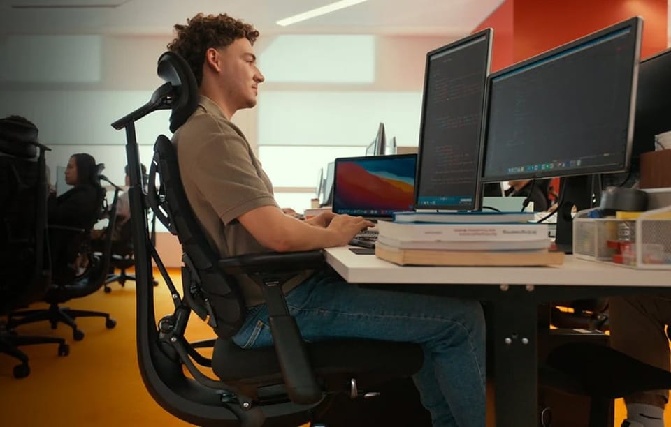 Mężczyzna siedzący na ergonomicznym fotelu biurowym z zagłówkiem pracuje przy biurku z dwoma monitorami w biurze