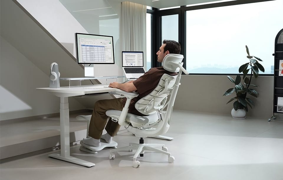Mężczyzna w brązowym t-shircie siedzi w białym ergonomicznym krześle przy białym biurku z monitorem