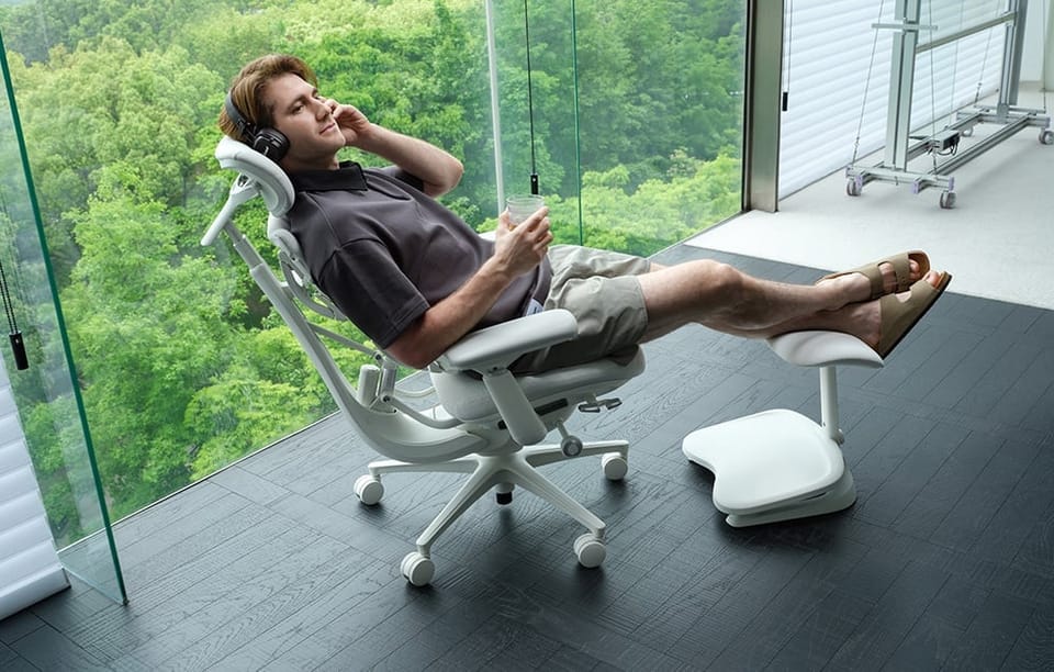 Mężczyzna w słuchawkach odpoczywa na białym fotelu ergonomicznym z podnóżkiem, trzymając szklankę napoju