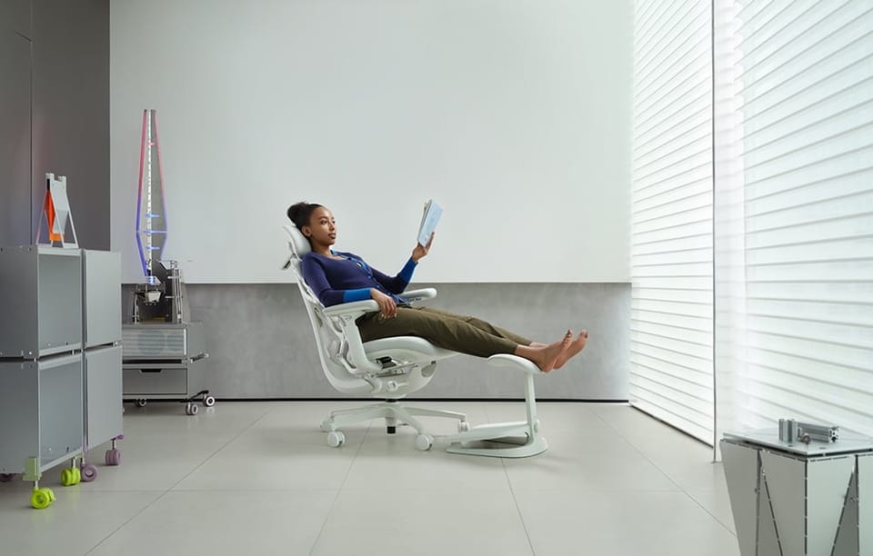 Kobieta w fioletowym swetrze czyta książkę na białym krześle ergonomicznym z podnóżkiem w jasnym pomieszczeniu
