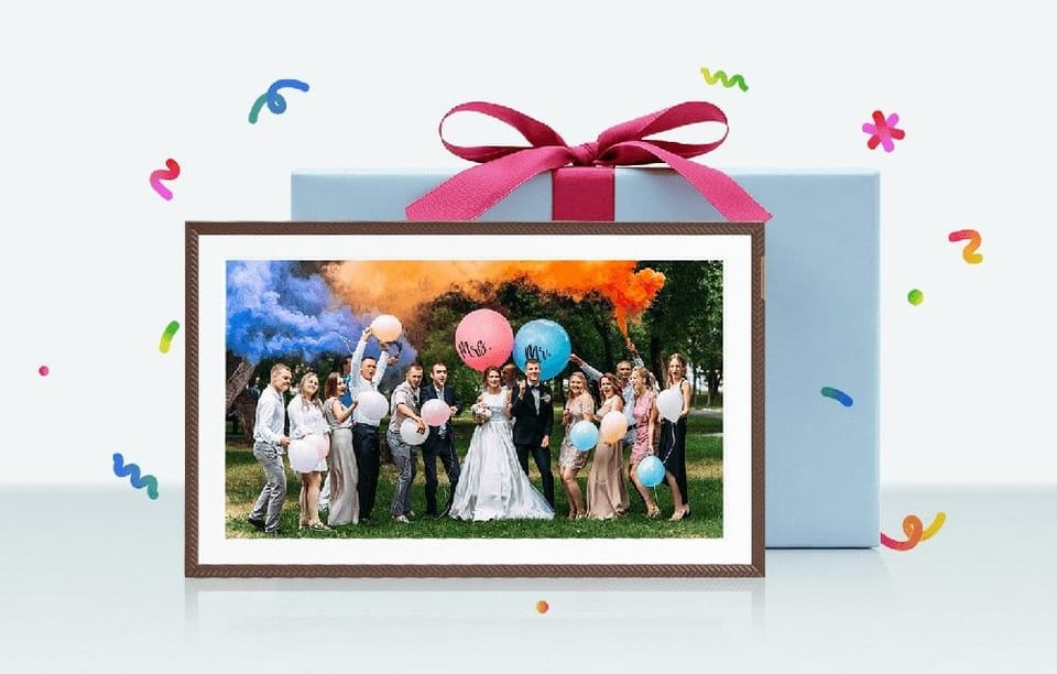 Cyfrowa ramka z fotografią grupy weselnej z balonami i dymem kolorowym, ustawiona przed zapakowanym prezentem