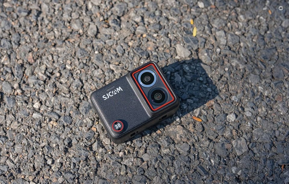 Kamera sportowa SJCAM z dwoma obiektywami i czerwoną obwódką leżąca na asfalcie.