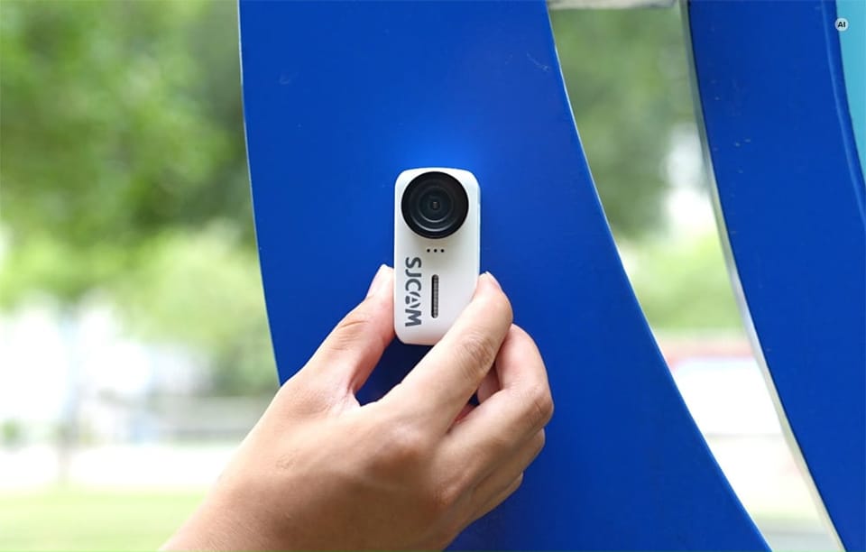 Dłoń przyczepiająca małą, białą kamerę sportową SJCAM do niebieskiej powierzchni na zewnątrz.