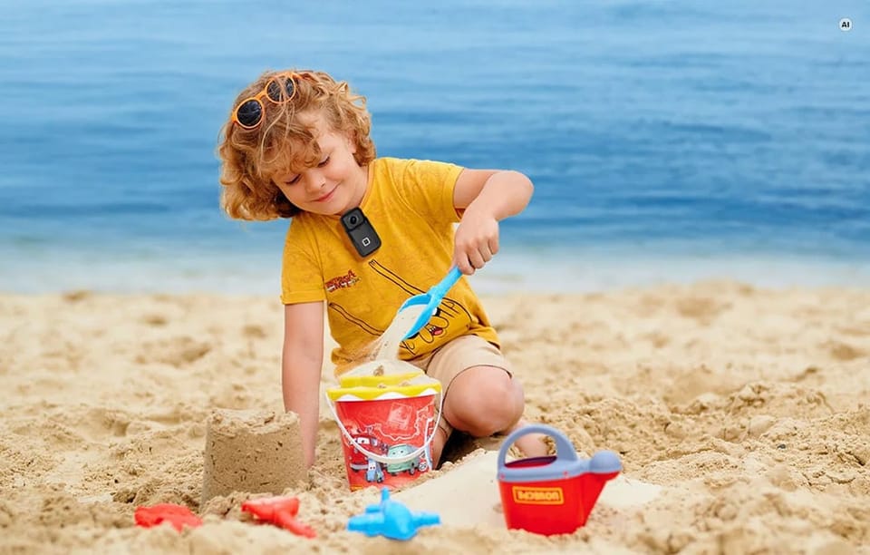Dziecko bawiące się zabawkami na piaszczystej plaży z przypiętą do koszulki małą kamerą.