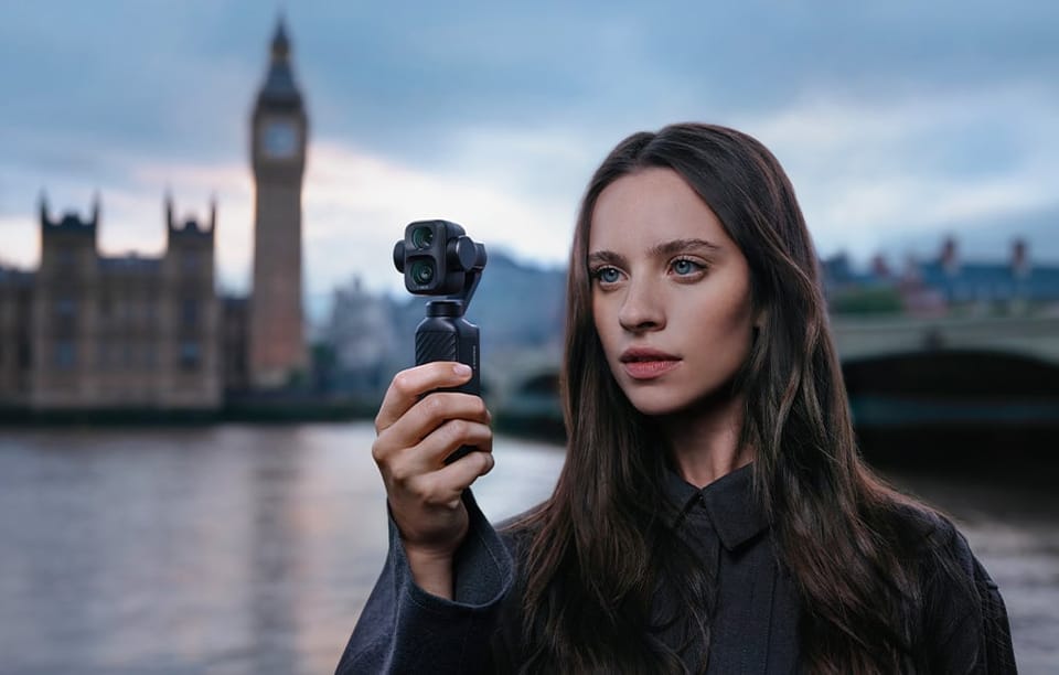 Młoda kobieta trzyma małą kamerę, stojąc nad rzeką w Londynie, z wieżą Big Ben w tle