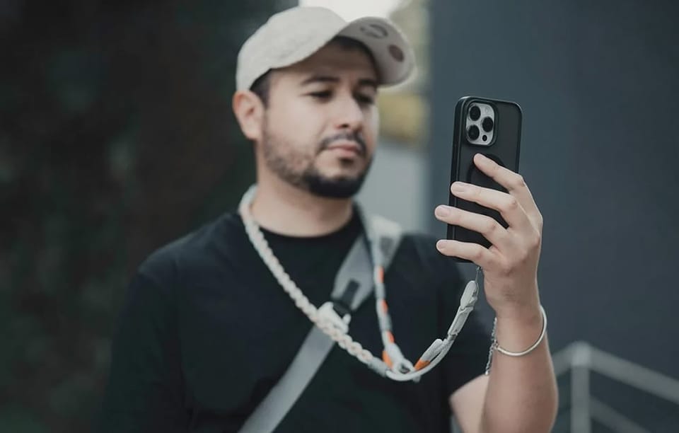 Mężczyzna w czapce z daszkiem patrzy na telefon w etui, który jest przypięty do jasnej, plecionej smyczy na jego szyi.